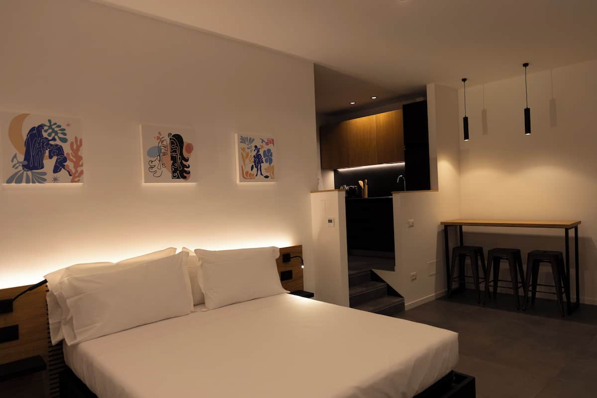 A modern hotel room with a bed, artwork on the wall, a desk with stools, and a kitchen area.