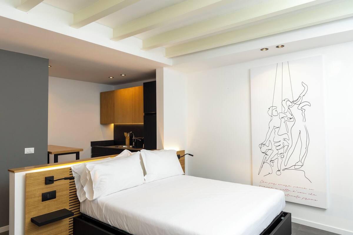 A modern bedroom with a bed, bedside tables, a wooden headboard, a wall-mounted artwork, and a kitchen area in the background.