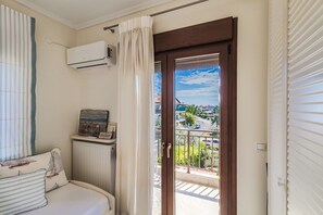 A bedroom with a bed, an air conditioner, a radiator, a window with curtains, and a balcony with a view of buildings and greenery. - Fibes Luxury Living 2 by Rentals Pro - Mouries Halkidiki (Mouries)