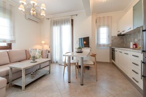 A modern living room with a sofa, a dining table with chairs, a TV, and a kitchen area with cabinets and a refrigerator. - Fibes Luxury Living 2 by Rentals Pro - Mouries Halkidiki (Mouries)