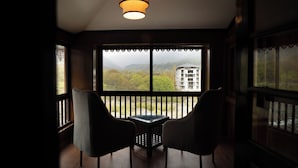 Habitación con vistas a un edificio y montañas a través de amplios ventanales. Hay dos sillas y una mesa en la habitación.