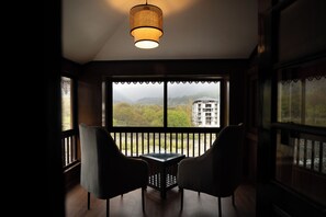 Habitación con vistas a un edificio y montañas a través de amplios ventanales. Hay dos sillas y una mesa en la habitación.