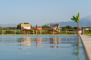 Área ao lado da piscina com duas cadeiras de vime, uma mesa com garrafa e copos, e um vaso de planta.