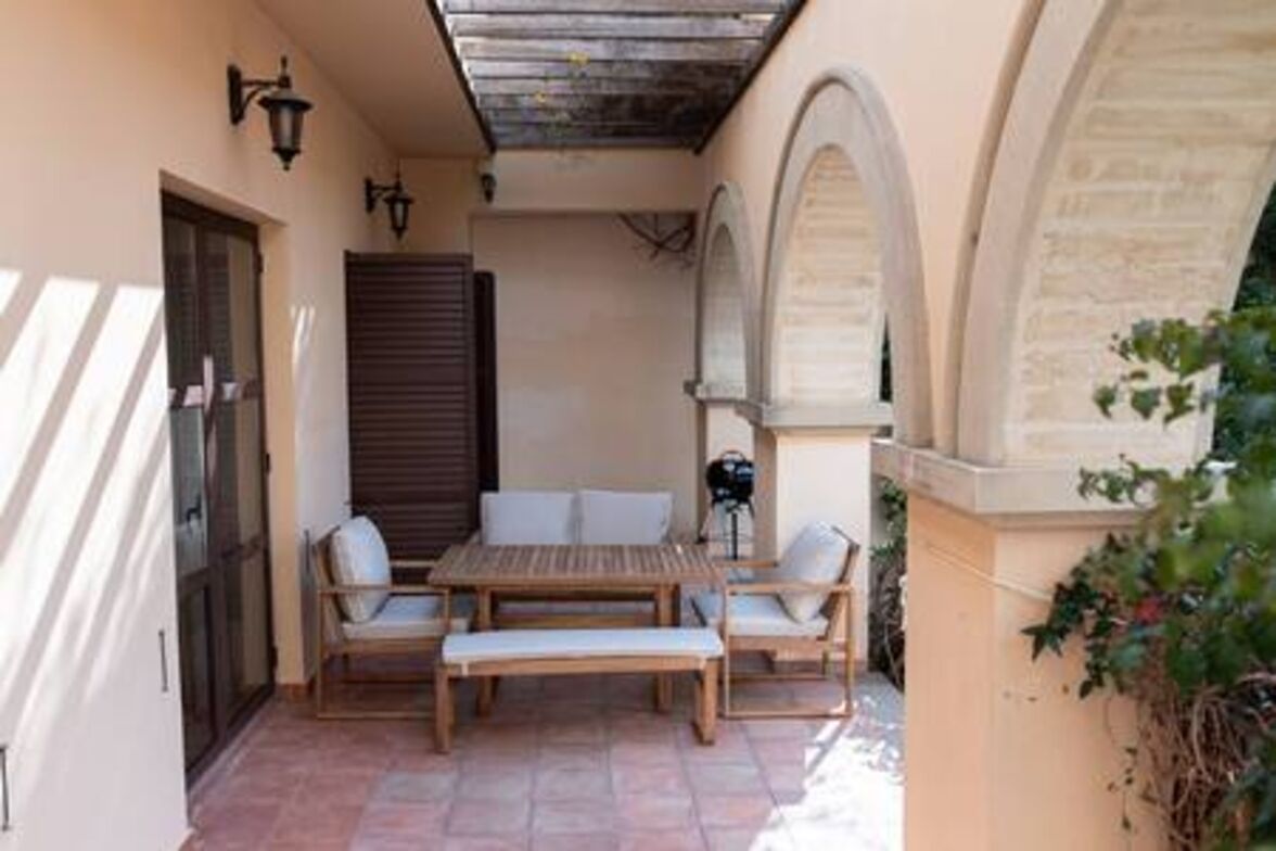 Una terrazza esterna con tavolo e sedie in legno, un pergolato sopra e un muro in pietra con archi.