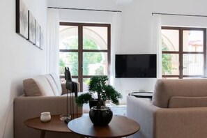 Ein Wohnzimmer mit einer Couch, einem kleinen Tisch mit Pflanze, einem Fernseher und großen Fenstern.