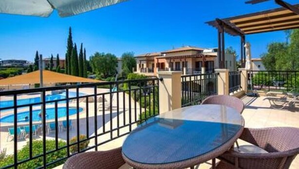 A balcony with a glass table and wicker chairs, overlooking a swimming pool and a building with a tiled roof. - BF12 Adjacent to Aphrodite Hills - Pool & Terrace (Kouklia)