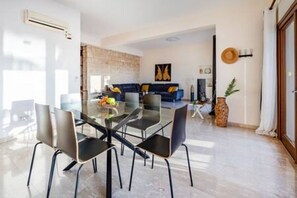 A modern dining area with a glass table and chairs, a living room with a sofa and decorative items, and a stone wall. - HG07 Adjacent to Aphrodite Hill - Pool & Terrace (Kouklia)