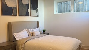 Un dormitorio con cama, mesita de noche, lámpara de pared, dos cuadros abstractos, ventana con persianas y un respiradero en el techo.