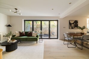 A modern living room with a green sofa, a dining table with chairs, a black coffee table, and a large sliding glass door leading to an outdoor area. - Sage Tones with Private Patio (London)