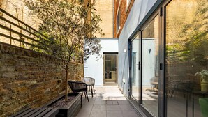 A narrow outdoor patio with a brick wall, a wooden bench, and a glass door leading to an interior space. - Sage Tones with Private Patio (London)