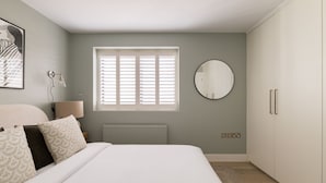 A bedroom with a bed, a round mirror, a window with blinds, a radiator, and a wardrobe. - Sage Tones with Private Patio (London)