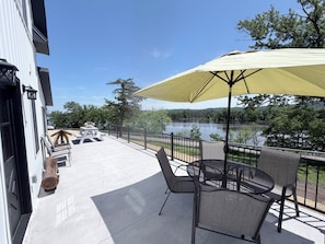 A patio with outdoor furniture, a table, and an umbrella, overlooking a lake and surrounded by trees.