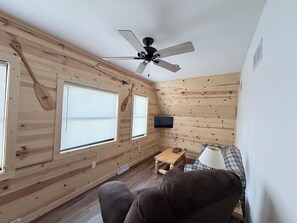A small, cozy room with wooden walls, a ceiling fan, a television mounted on the wall, a wooden table, and a couch.