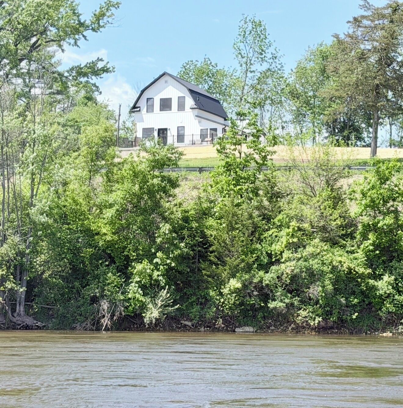 A white house with a black roof, surrounded by dense green trees and a body of water.