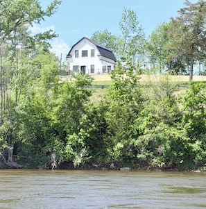 A white house with a dark roof, surrounded by dense green foliage and a body of water in the foreground.