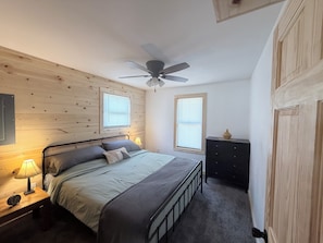 Un dormitorio con cama, lámpara de noche, ventilador de techo, dos ventanas y cómoda.