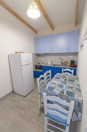 Eine kleine Küche mit Kühlschrank, Herd, Spüle und blauen Schränken. Ein Esstisch mit Stühlen und einer Tischdecke mit Muster ist ebenfalls vorhanden.