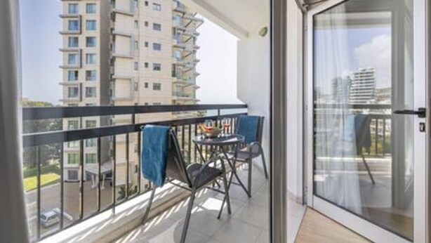 Un balcon avec une table et des chaises, une vue sur un immeuble de grande hauteur et une porte vitrée.