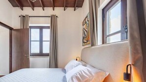 Um quarto com cama, janela, cortinas, uma luminária na parede e um quadro.