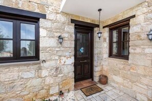 A stone exterior with a dark wooden door, two windows with dark frames, and a hanging light fixture. - Stroumpi Private Pool Villa (Stroumpi)