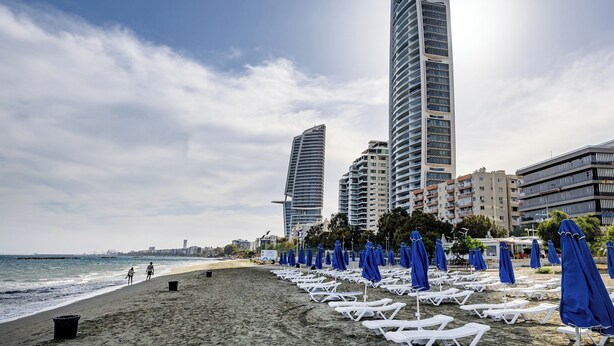 Une plage avec des chaises longues blanches et des parasols bleus, de grands immeubles en arrière-plan et l’océan.