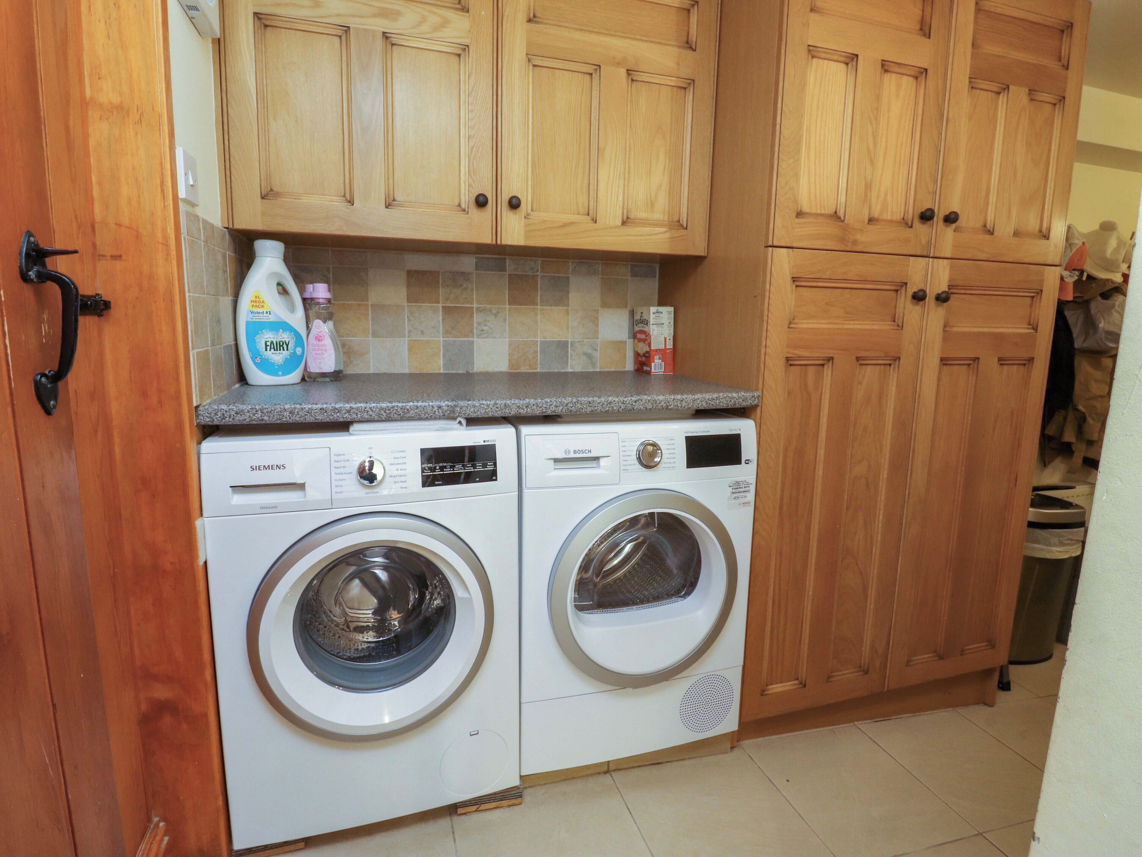 A laundry room with two front-loading washing machines, wooden cabinets, and a tiled backsplash.