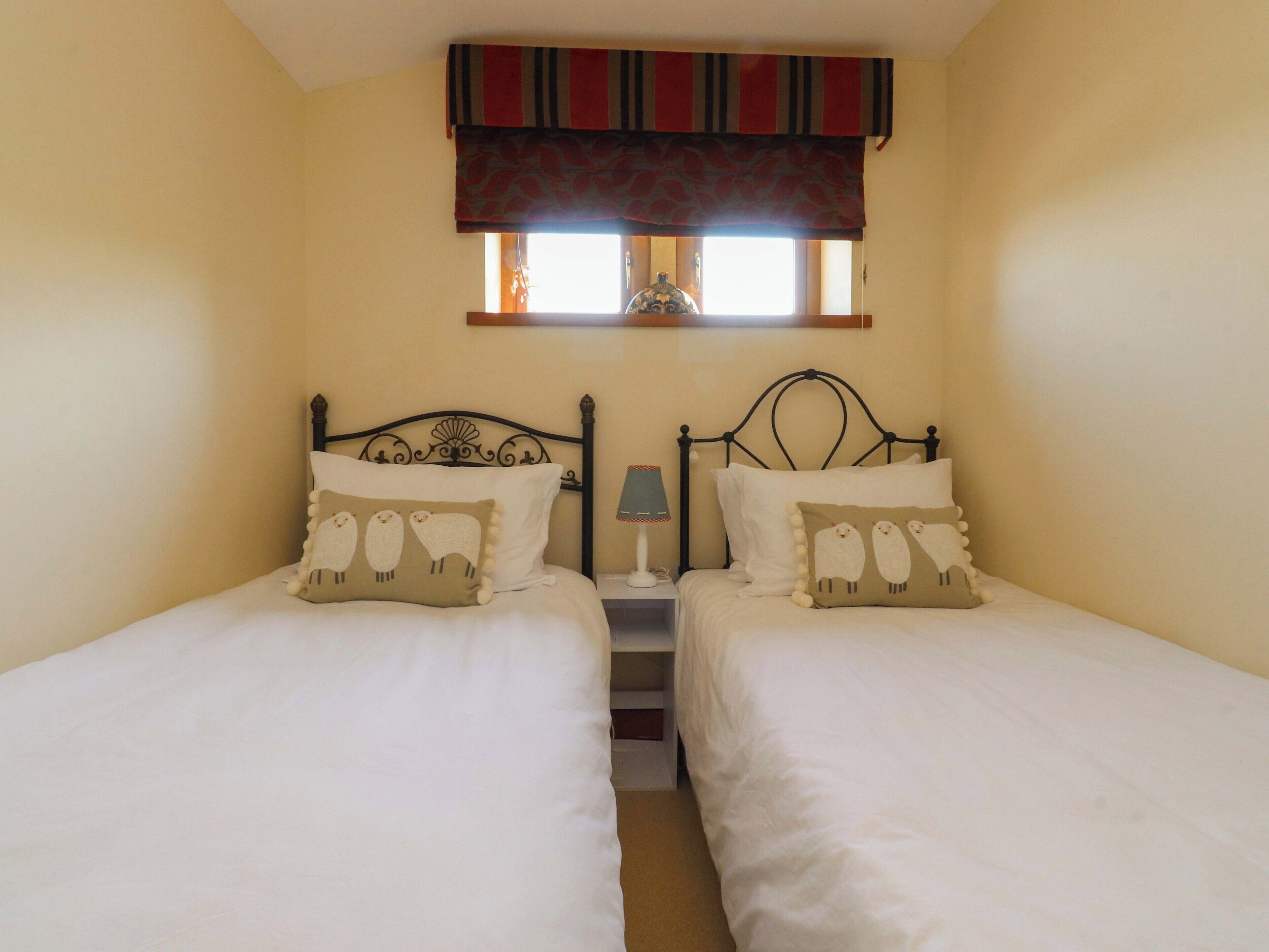 Una habitación con dos camas, cada una con almohadas a juego, una ventana con un visillo a rayas y una mesita de noche con una lámpara.