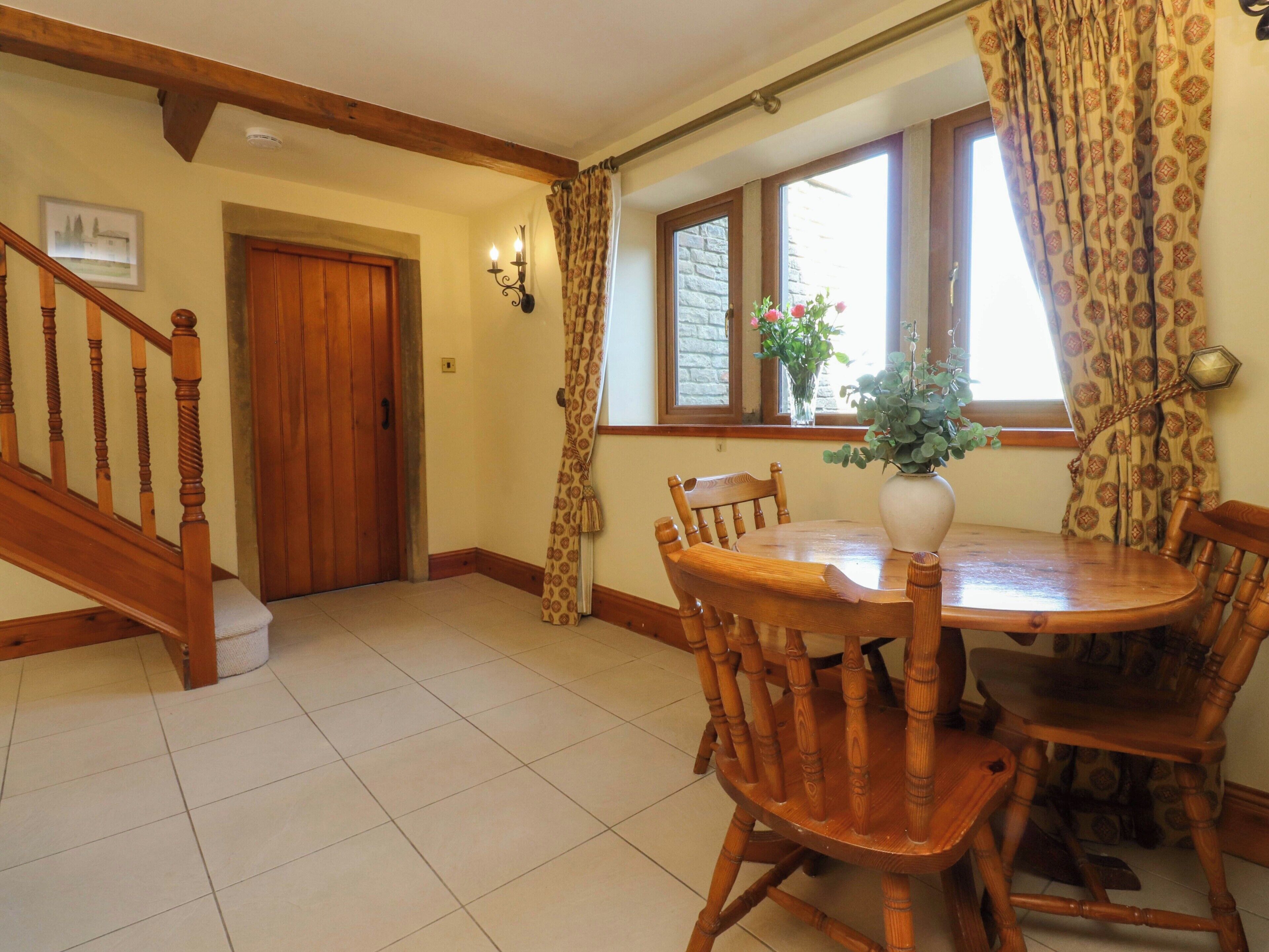 A dining area with a wooden table and chairs, a vase with flowers, a window with patterned curtains, and a wooden staircase.