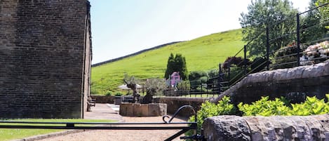 A stone building with a gravel courtyard, a metal gate, and a green hill in the background.
