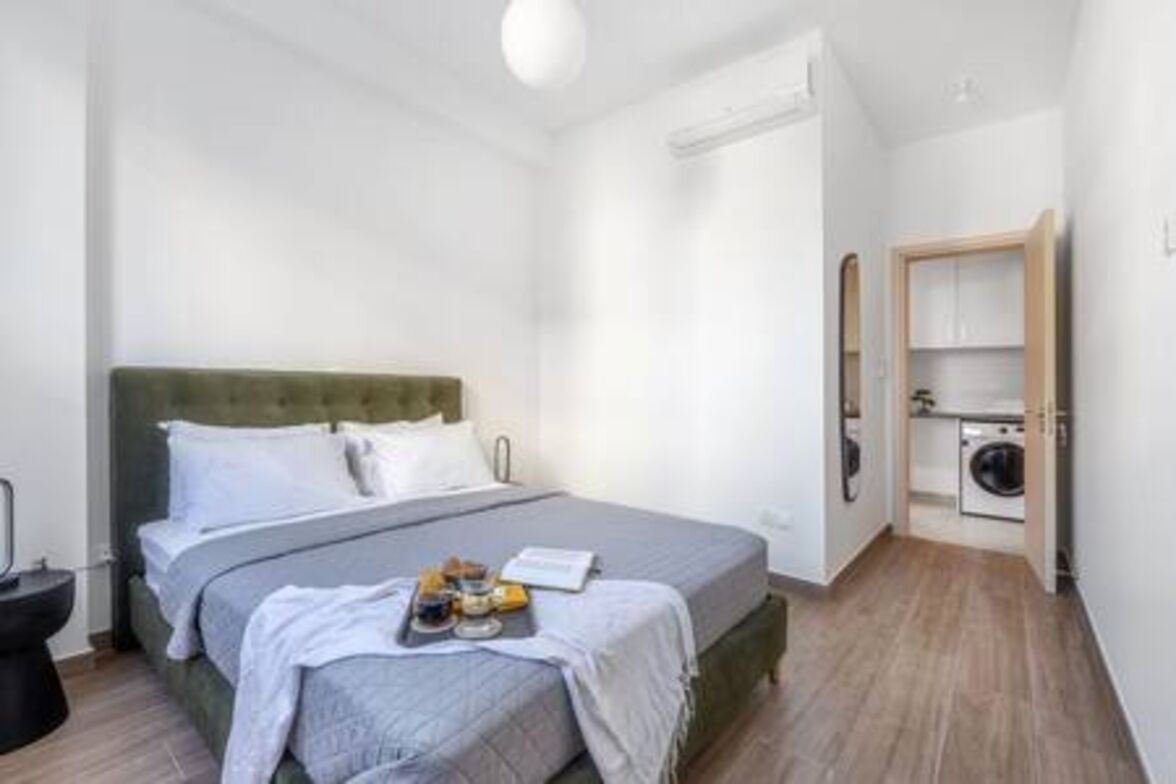 A small, modern bedroom with a bed, a bedside table, a nightstand, a wall-mounted heater, and a laundry area with a washing machine.