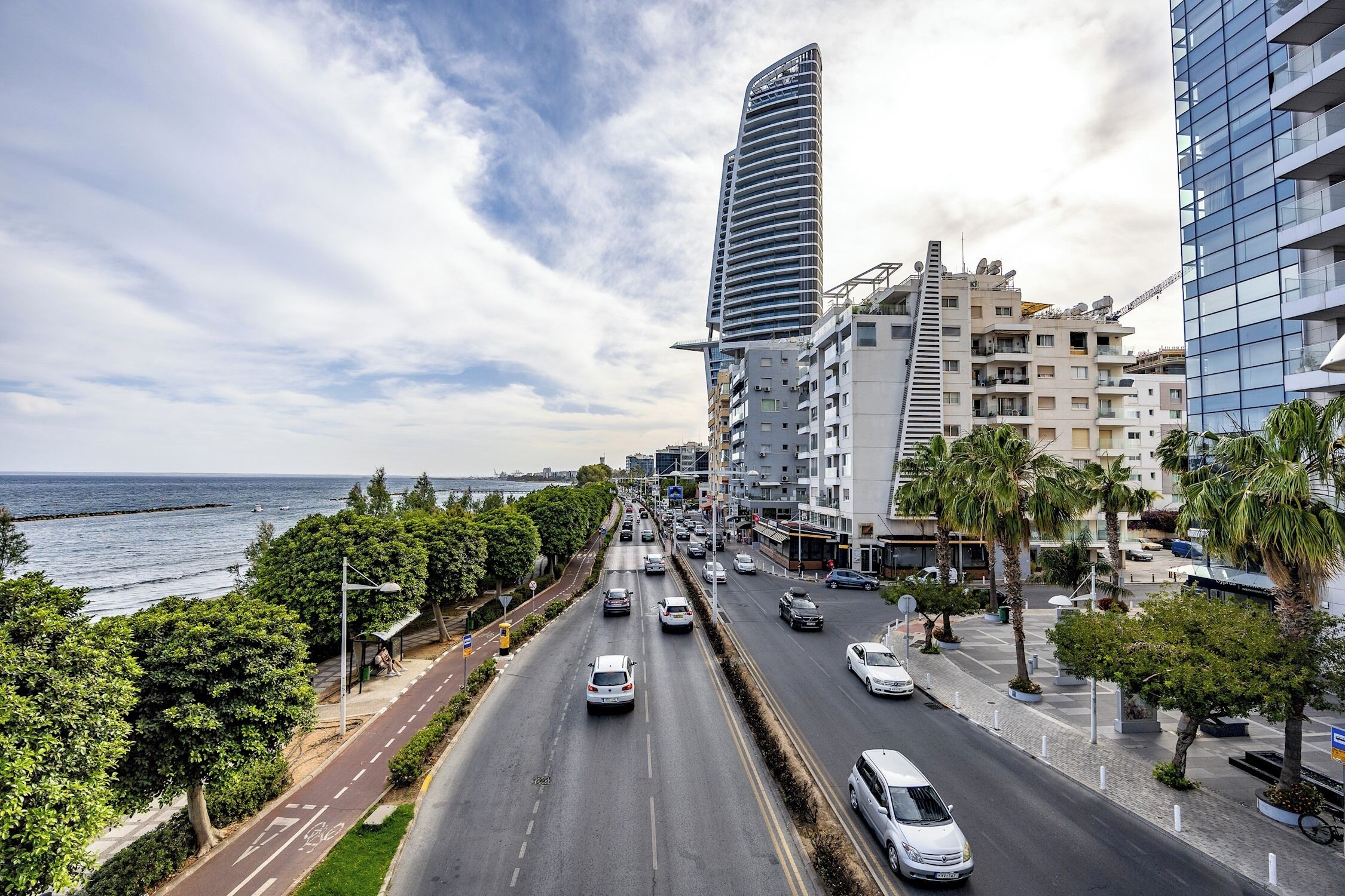 Paisagem costeira com edifícios modernos, uma estrada com veículos e um passeio para pedestres à beira-mar.