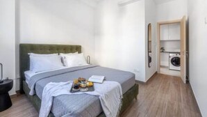 Ein kleines, modernes Schlafzimmer mit einem Bett, einem Nachttisch, einer Wandheizung und einem Waschbereich mit Waschmaschine.