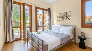 Quarto com estrutura de cama em metal, mesa de cabeceira, janela com vista para edifícios e porta que dá para área externa.
