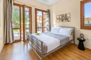 Ein Schlafzimmer mit Metallbettgestell, einem Nachttisch, einem Fenster mit Blick auf Gebäude und einer Tür zu einem Außenbereich.