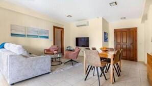 Sala de estar com um sofá, uma mesa de centro e uma TV. Há uma área de jantar com uma mesa e cadeiras.