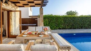 Uma área de piscina ao ar livre com mesa e cadeiras, um espaço de estar coberto e um conjunto de sebe ao fundo.