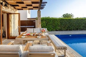 Ein Poolbereich im Freien mit Tisch und Stühlen, einer überdachten Sitzgelegenheit und einer Hecke im Hintergrund.