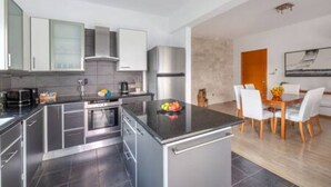 Uma cozinha moderna com eletrodomésticos de aço inoxidável, um espaço de refeições com mesa de madeira e cadeiras brancas, e vista para o exterior.