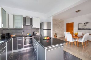 A modern kitchen with stainless steel appliances, a dining area with a wooden table and white chairs, and a view of the outdoors. - Lena Court - Spacious 3bdr, City Views (Limassol)