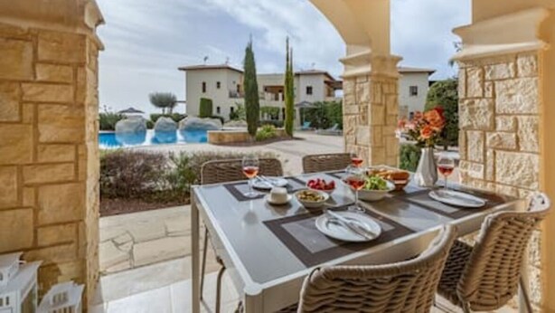 An outdoor dining area with a table set for two, stone pillars, and a view of a swimming pool and buildings in the background. - BK02 Adjacent to Aphrodite Hills – Pool & Sea View (Kouklia)
