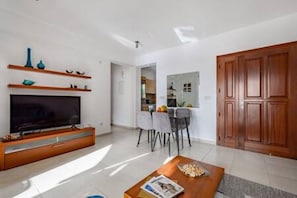 A modern living room with a flat-screen TV, wooden entertainment unit, dining area with chairs, and a large wooden door. - BK02 Adjacent to Aphrodite Hills – Pool & Sea View (Kouklia)