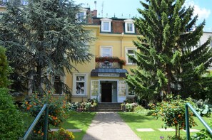 A yellow building with a balcony, a door, and a garden with flowers and trees. - defaultName (Wien)