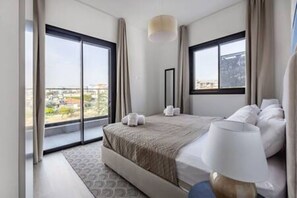 A hotel room with a large bed, a bedside table, a lamp, and a view of the city through large windows.