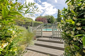 A wooden deck leading to a gated pool area surrounded by lush greenery and a clear sky.