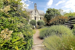 Un bâtiment en pierre doté d’une tour centrale, entouré d’une végétation luxuriante et d’un chemin de gravier menant à l’entrée.