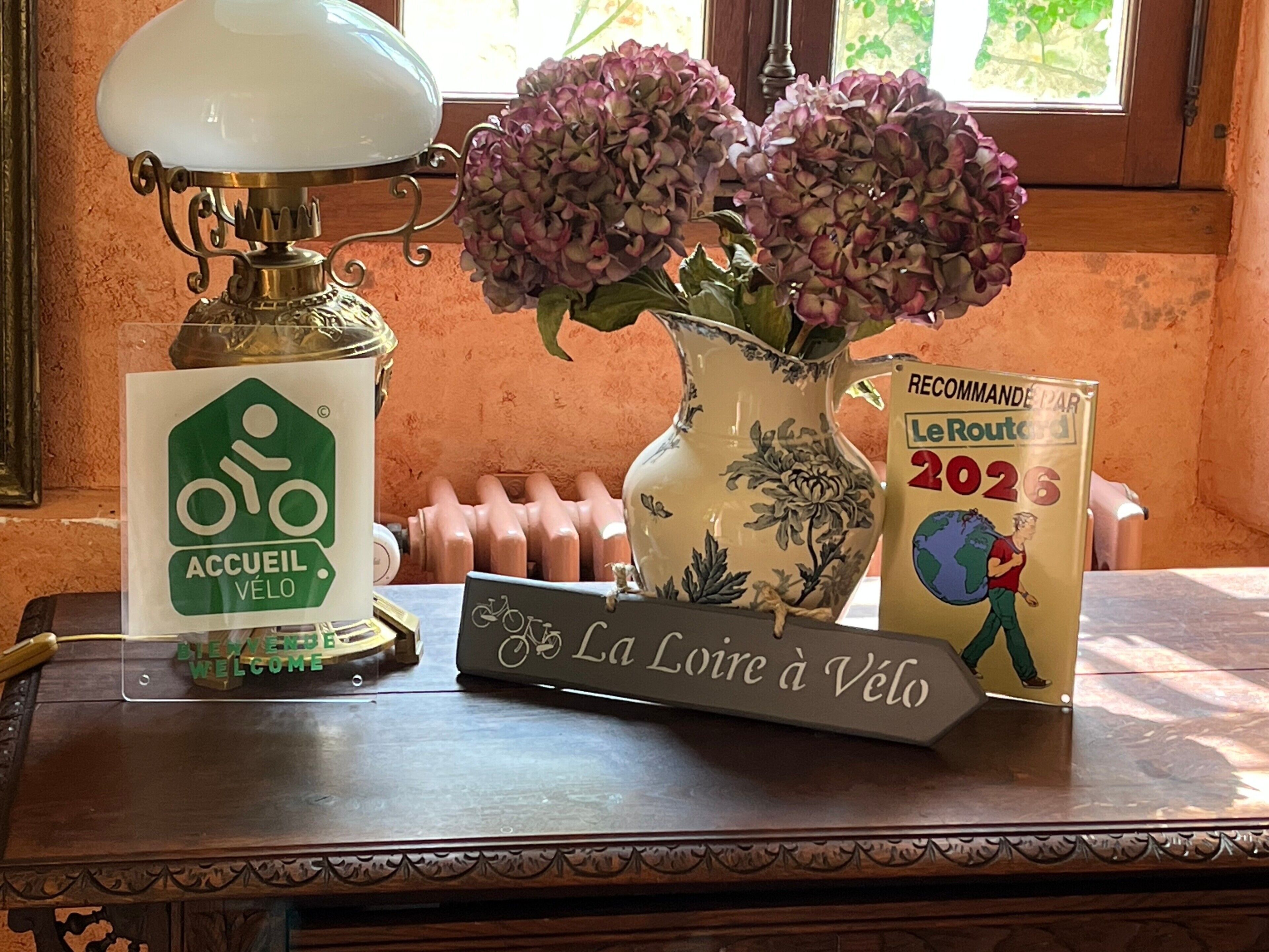 A table with a bicycle sign, a welcome sign, a decorative vase with flowers, and a sign for Le Route 2026.