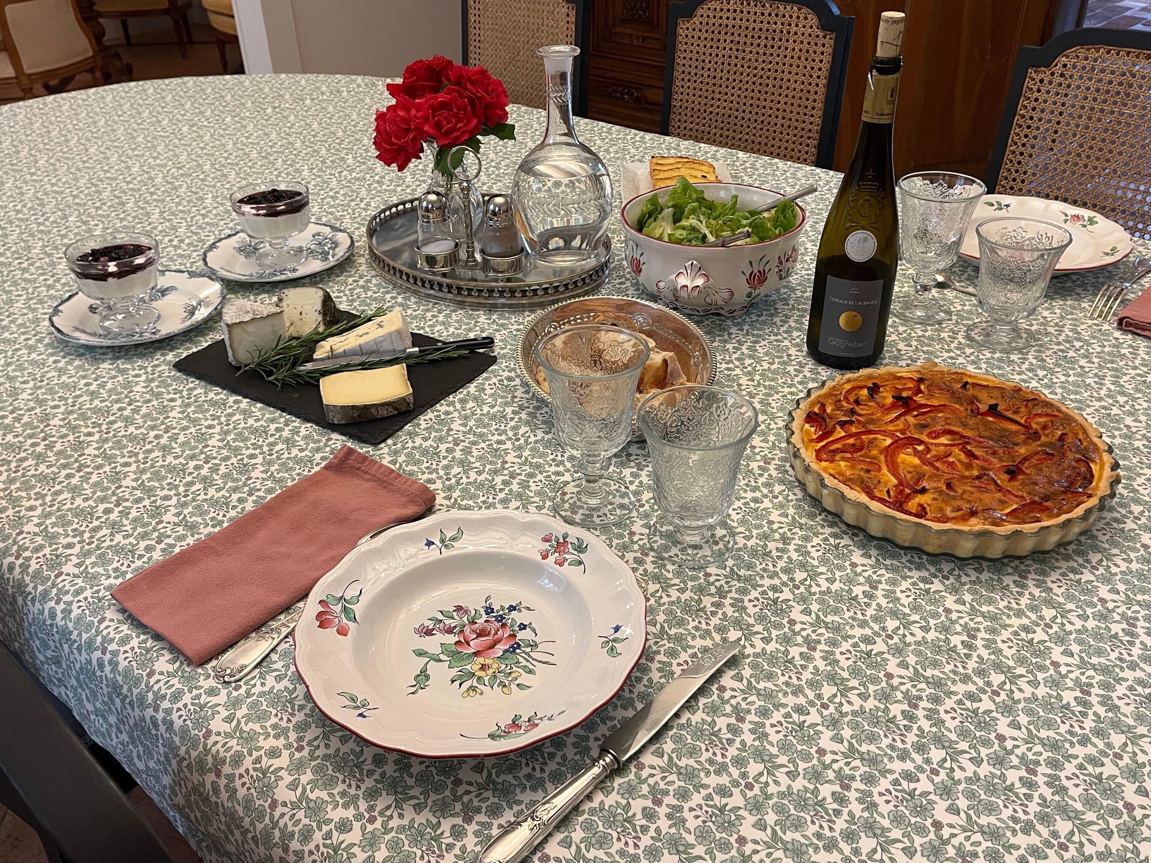 Una mesa para comer con mantel floreado, un pastel, una botella de vino, un tazón de ensalada y varios quesos y pan.