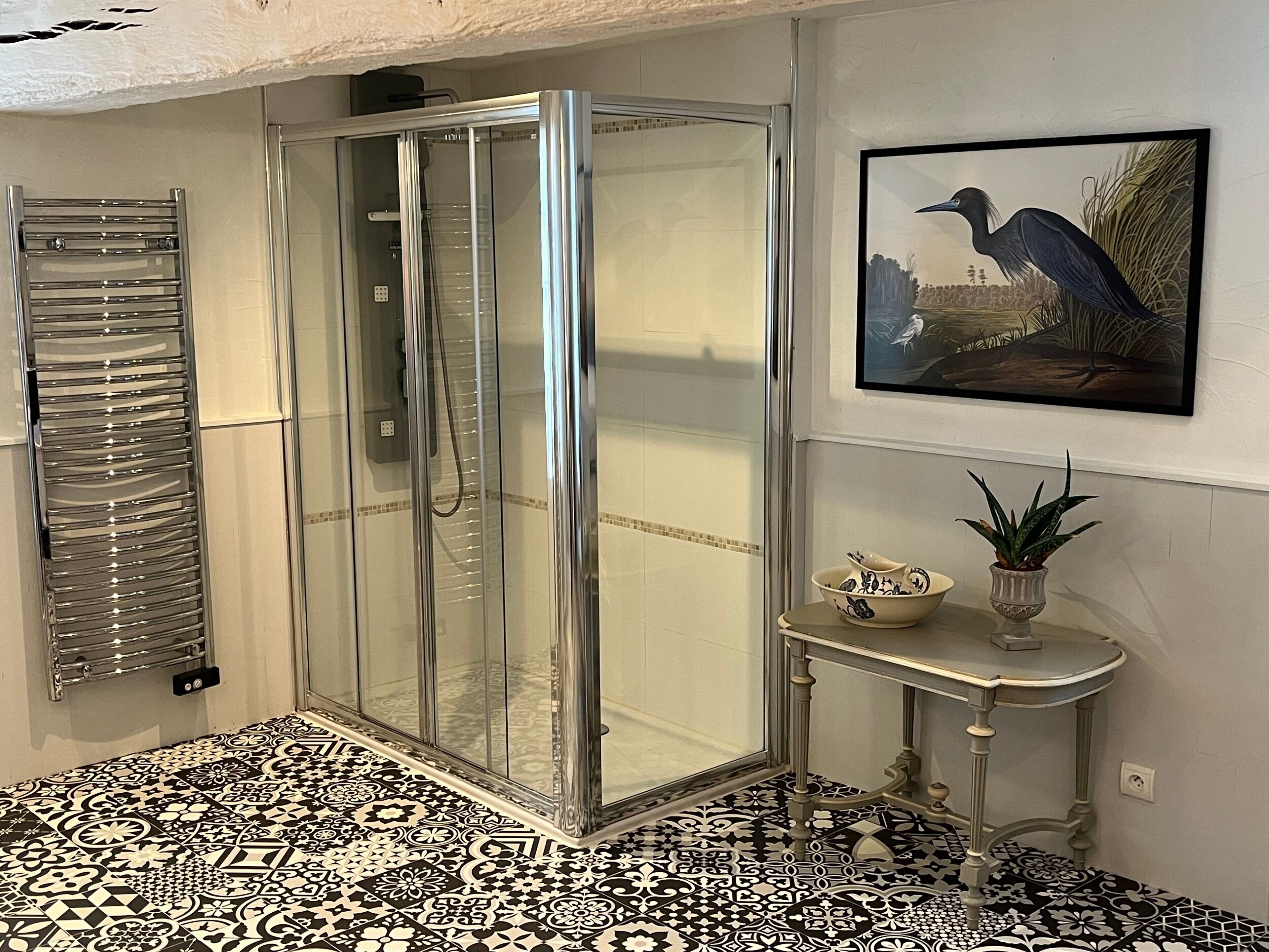 A bathroom with a glass shower enclosure, a tiled floor, a sink with a decorative bowl, and a framed picture on the wall.