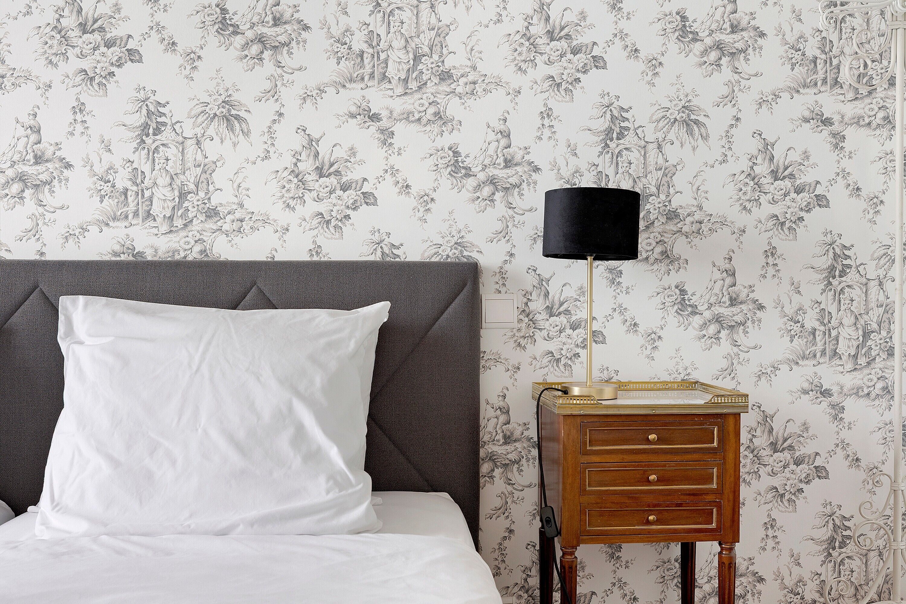 A bed with a white pillow and a grey headboard, a wooden nightstand with a lamp, and a floral wallpaper pattern.