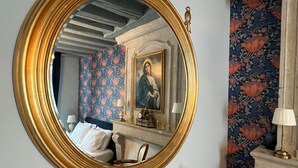 Een kamer met een grote, versierde spiegel, een bed, een kaptafel en een muur met bloemenbehang, plus een klein beeldje op de kaptafel.